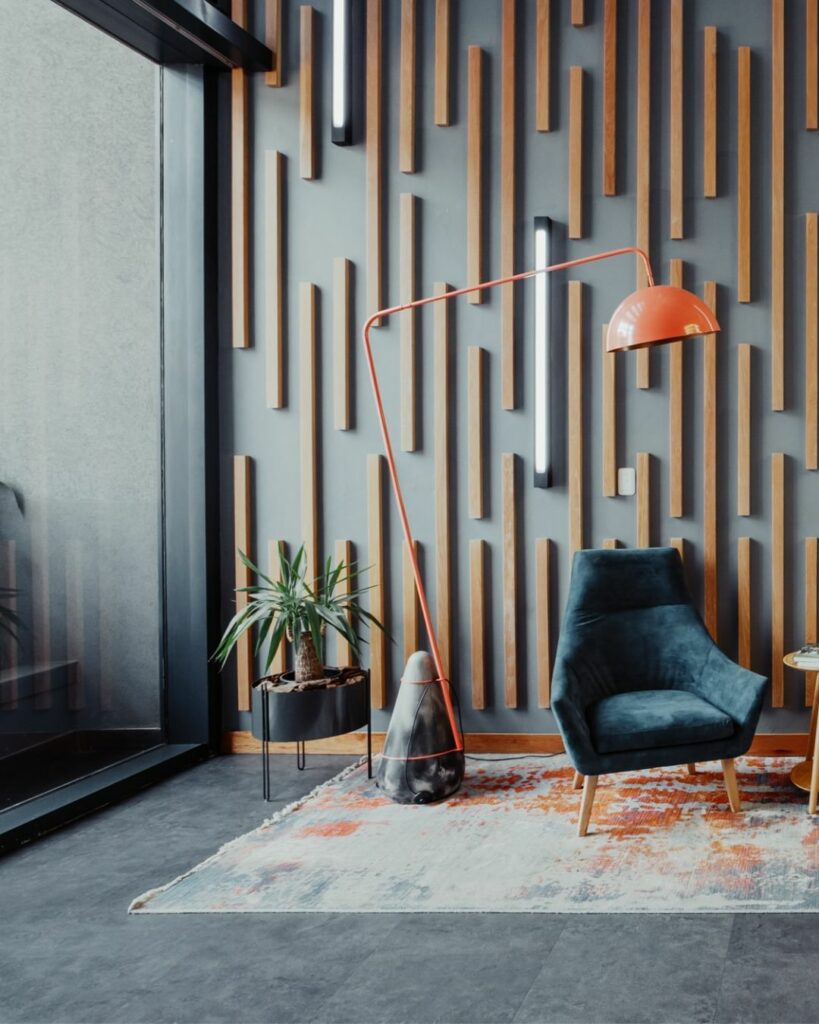 A modern seating area features two dark blue chairs, a small round side table with a book on it, and a tall orange floor lamp. The backdrop is a wall with vertical wooden slats and vertical strip lights. A colorful rug partially covers the floor.
