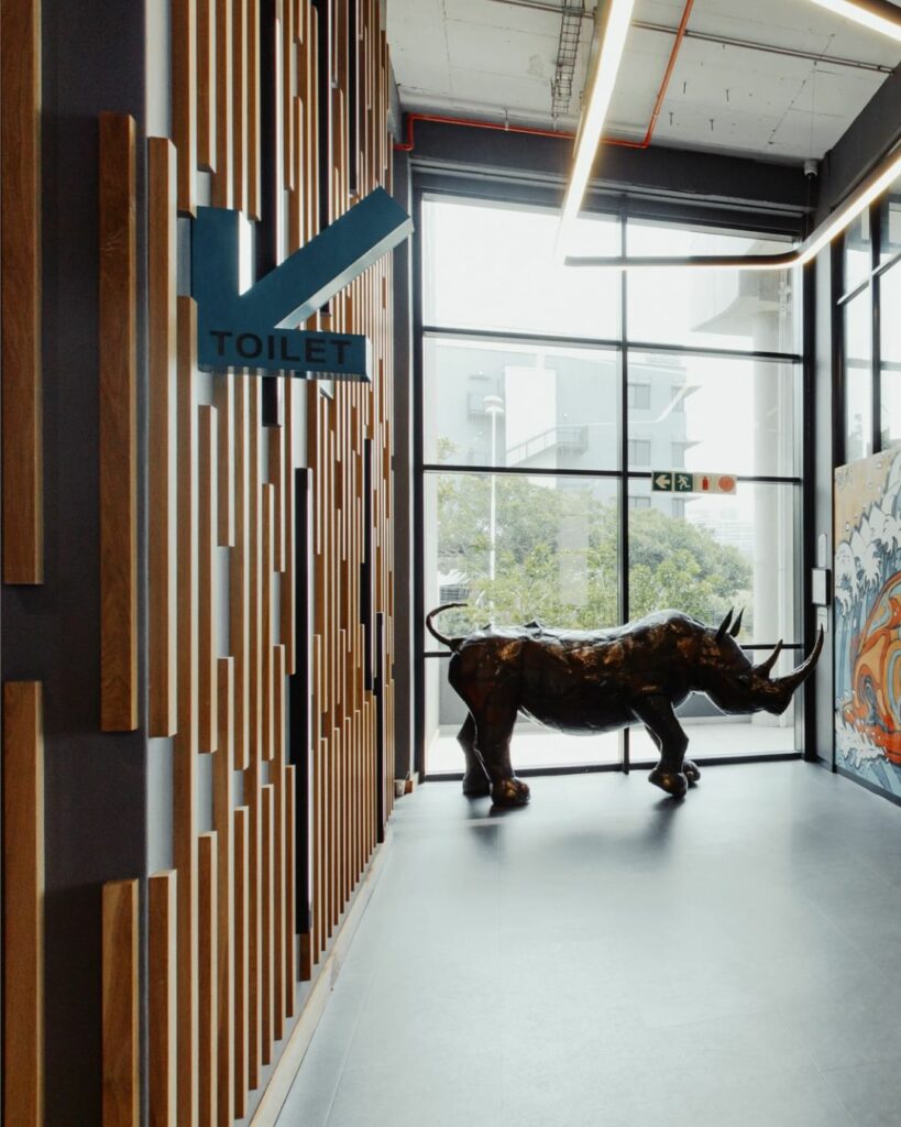 A modern hallway features a large decorative rhinoceros sculpture. On the left wall, there is a geometric wooden design with a blue "TOILET" sign. Large windows at the end of the hallway let in natural light, illuminating a vibrant mural on the right wall.