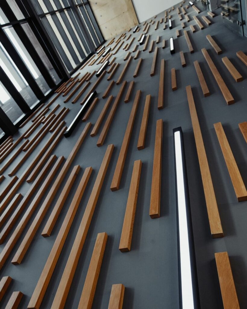 A modern wall design featuring horizontal wooden slats of varying lengths and slim LED lights, creating an abstract pattern. The wall extends vertically, with large windows on the left allowing natural light to enter the space.