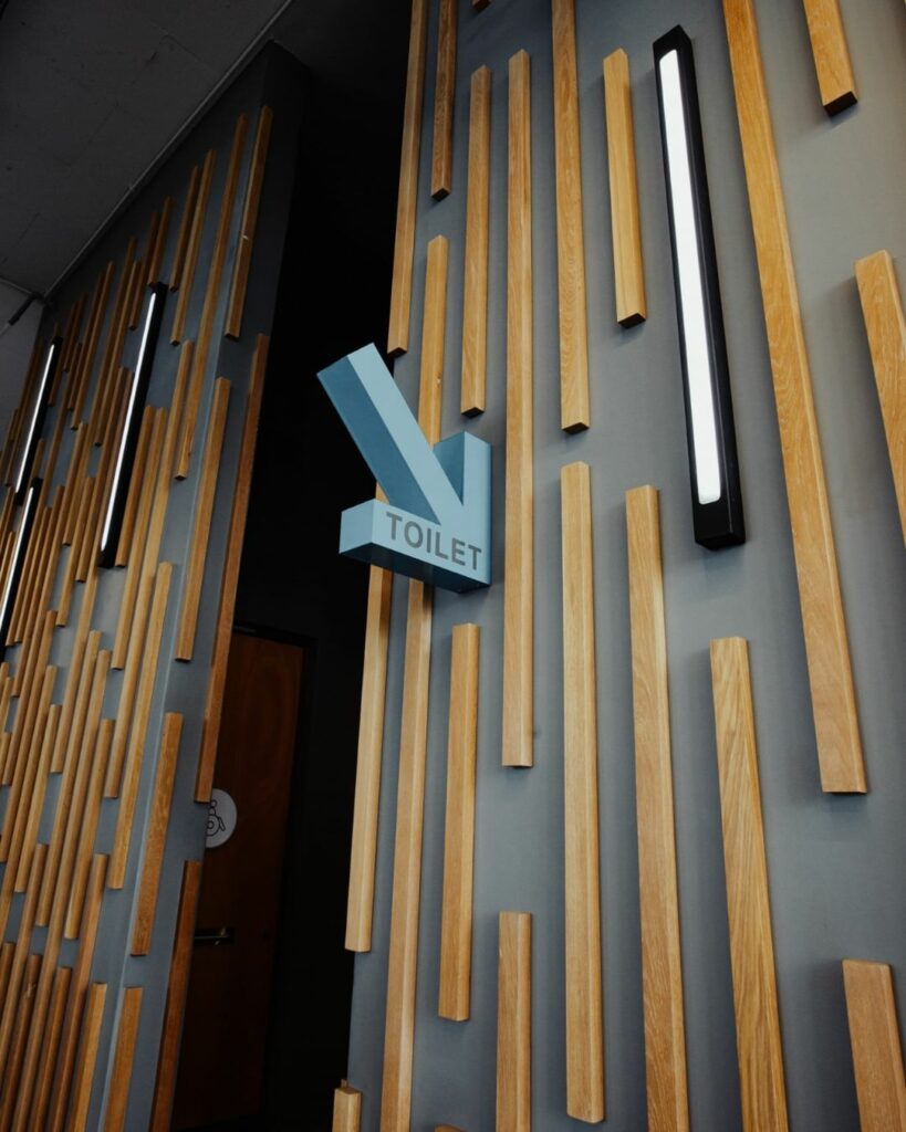 A modern wall features vertical wooden slats with a dark backdrop. Attached to the wall is a blue arrow-shaped sign pointing downward, labeled "TOILET." The wooden door beneath the sign has a restroom symbol on it. The area is well-lit.