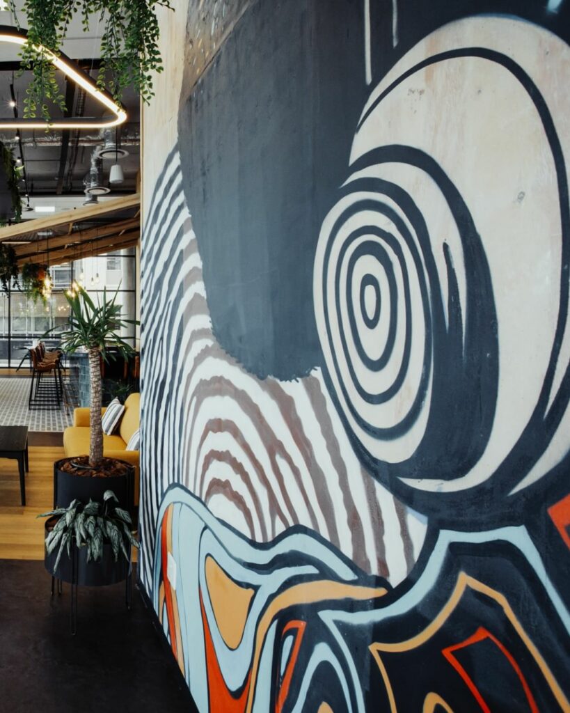 A modern office space with a large, abstract mural on one wall featuring swirling black and white patterns and warm colors. The area includes potted plants, a mustard yellow sofa, contemporary furniture, and industrial-style lighting. Natural light filters in through large windows.