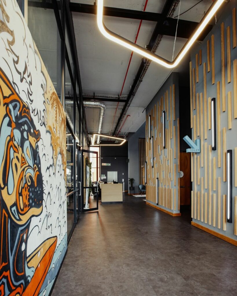A modern hallway with artistic wooden paneling on the right wall and a large, colorful mural on the left wall. Contemporary ceiling lights hang from a black ceiling frame, and at the end of the hallway, there is a glass-walled office space.