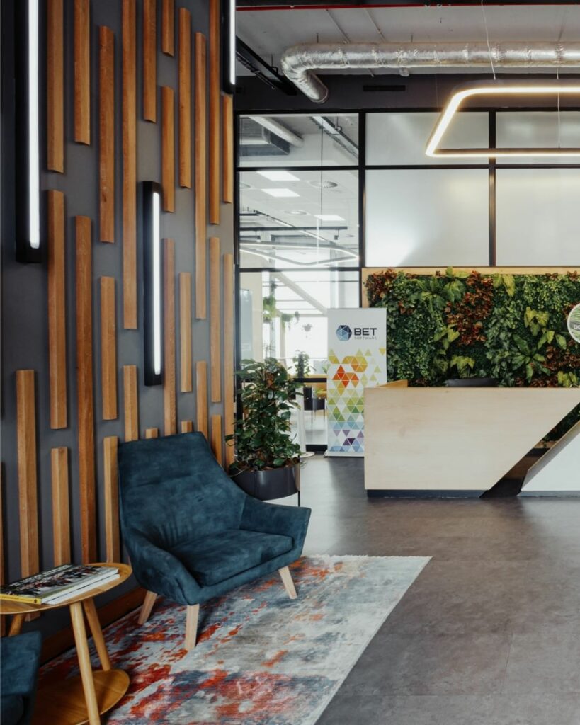 A modern office lobby with dark walls featuring vertical wooden slats, a comfortable blue chair on a patterned rug, and a small wooden round table with books. There is a reception desk in front of a glass partition adorned with a lush green plant wall.