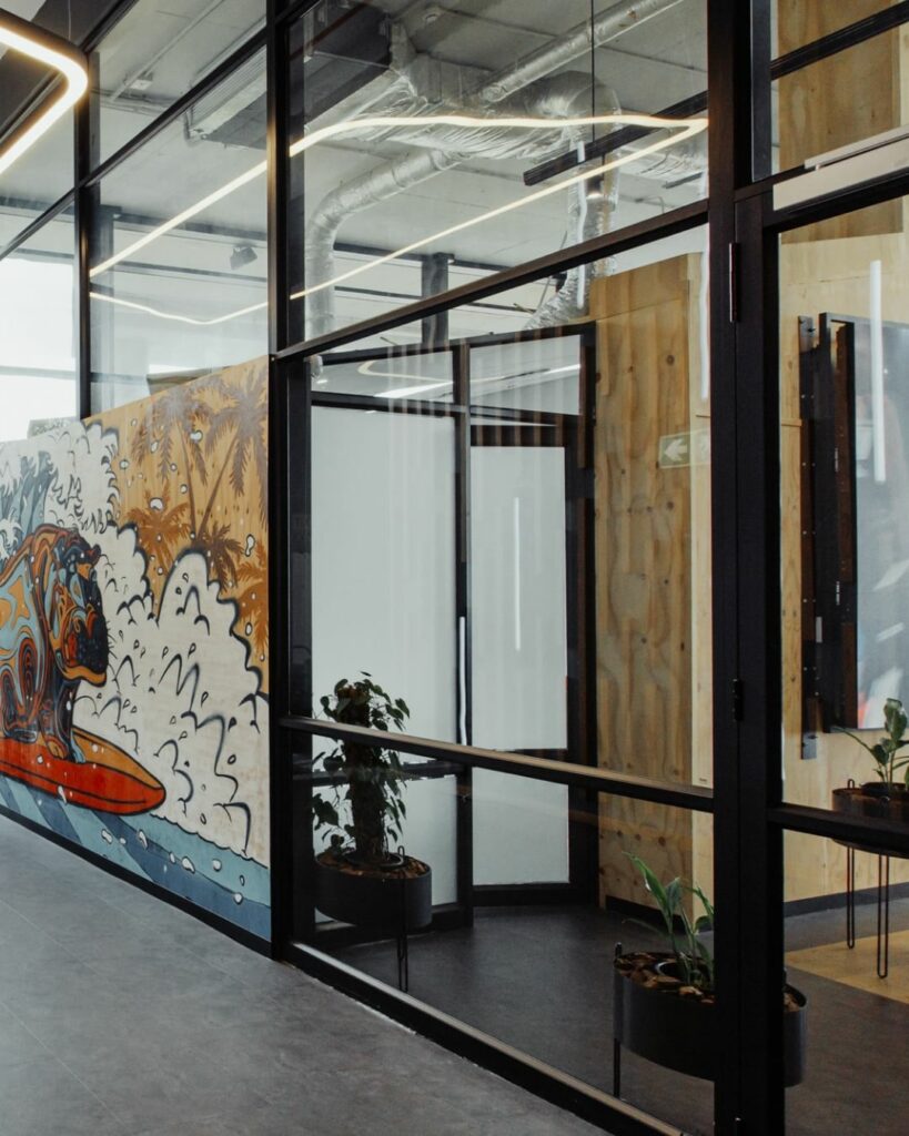 A modern office hallway with glass walls and metal frames. A vibrant mural of a colorful animal surfing on a wave is painted on the wall. Two potted plants sit near the entrance of an office with a wooden door. Industrial ceiling features are visible above.
