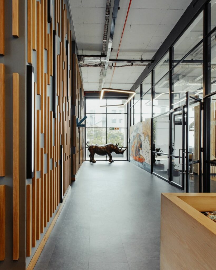 A modern hallway features a large decorative rhinoceros sculpture. On the left wall, there is a geometric wooden design with a blue "TOILET" sign. Large windows at the end of the hallway let in natural light, illuminating a vibrant mural on the right wall.