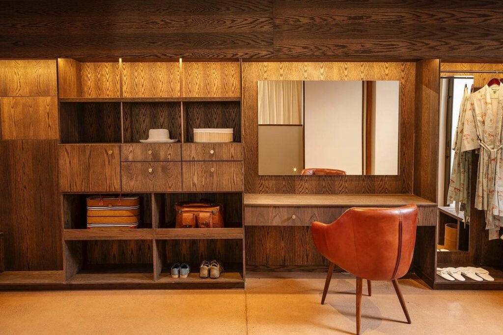 A stylish dressing room with dark wood shelves filled with clothes and accessories, featuring a large mirror, a desk, and a brown leather chair on a wooden floor.
