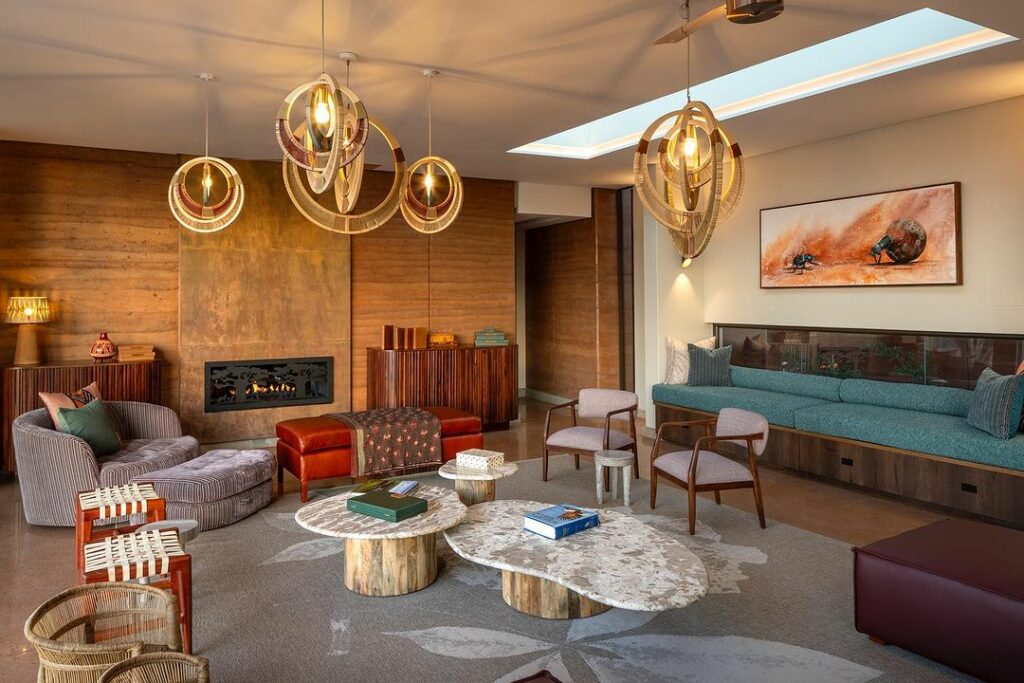 A stylish living room with modern rustic decor, featuring circular chandeliers, a fireplace, leather couches, and a glass wall overlooking a serene outdoor view.