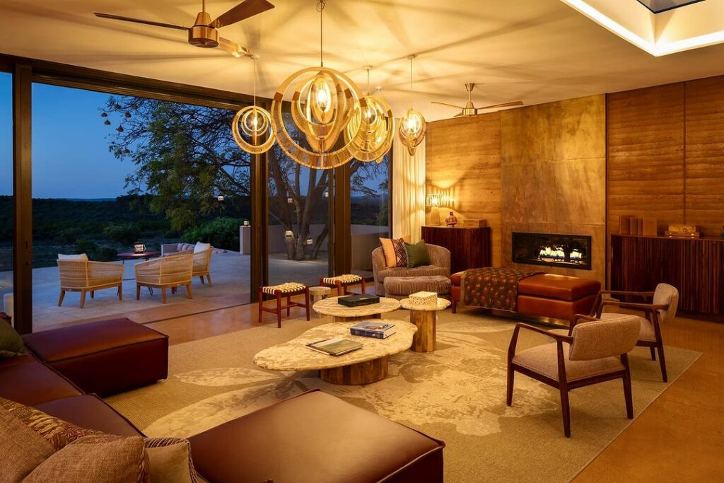 A stylish living room with modern rustic decor, featuring circular chandeliers, a fireplace, leather couches, and a glass wall overlooking a serene outdoor view.