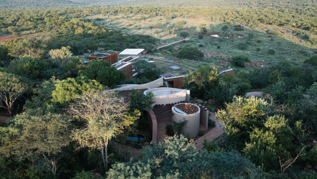 Aerial view of Melote House, a unique architectural complex with circular structures, nestled among dense greenery in a bush landscape. The buildings feature earth-toned walls and round roofs.