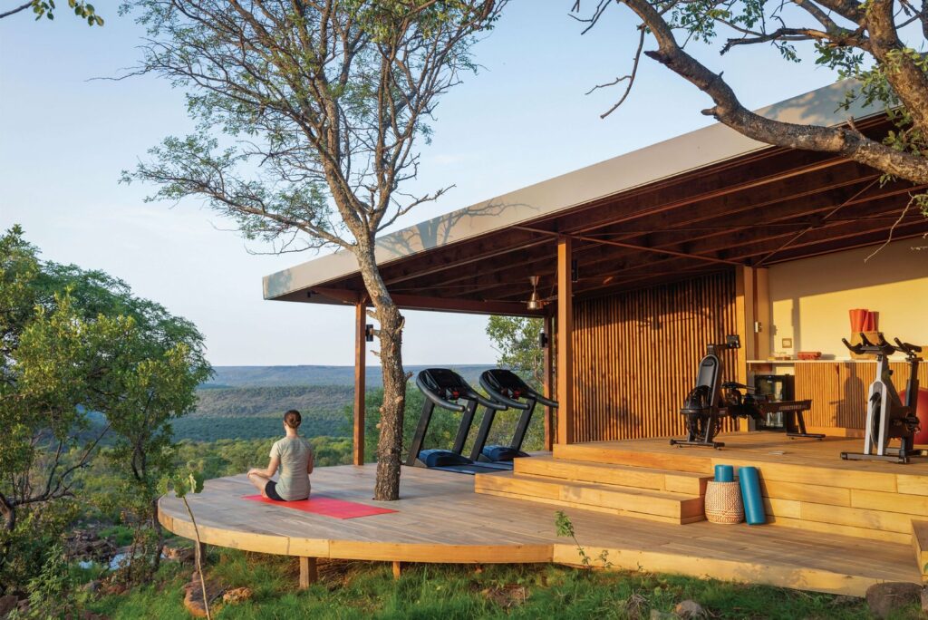 A woman meditates on a yoga mat on an outdoor platform at Melote House, overlooking a lush landscape, near a gym area with treadmills and wooden accents.