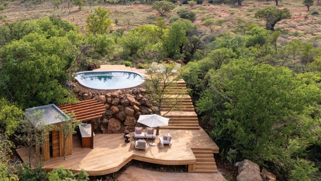 An aerial view of a luxurious outdoor pool at Melote House, featuring a wooden deck and seating area, surrounded by lush green trees and a rocky landscape.