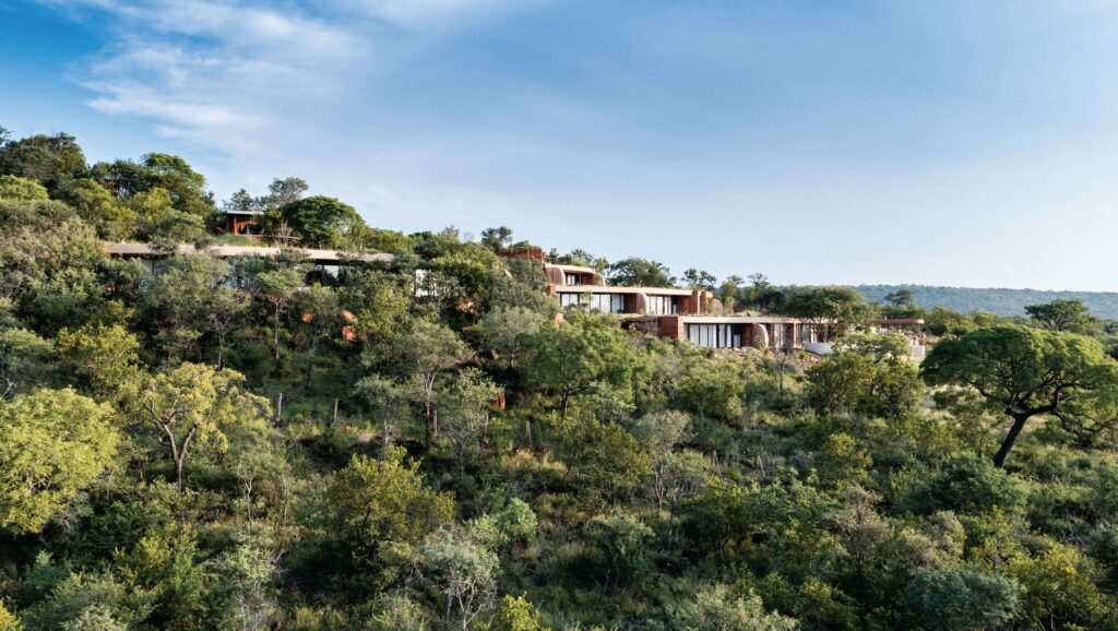 Wide view of a luxury safari lodge with an outdoor pool, featuring expansive terraces and abundant glazing, surrounded by lush vegetation.