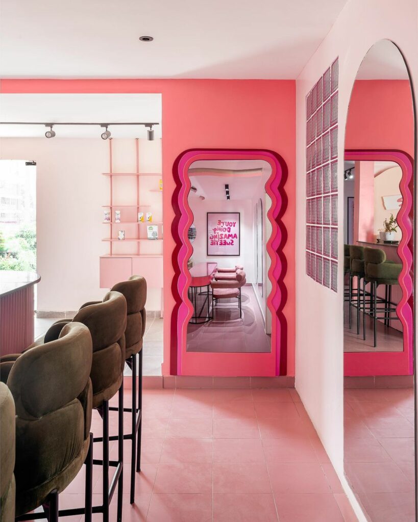 A vibrant room with a bar and chairs, featuring a pink and green color scheme and a mirror with a wavy frame.