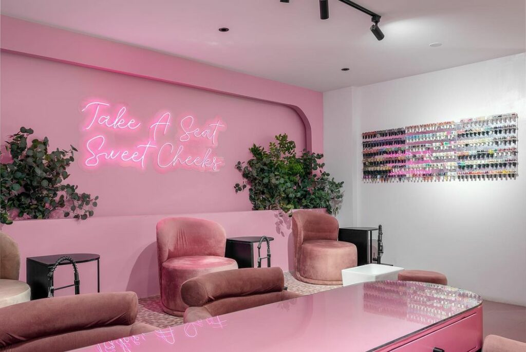 A pink salon interior with a neon sign that reads "Take a Seat Sweet Cheeks".