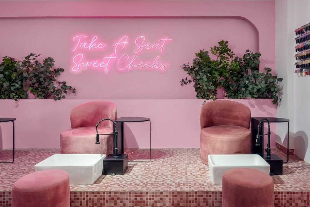 A pink salon interior with a neon sign that reads "Take a Seat Sweet Cheeks".