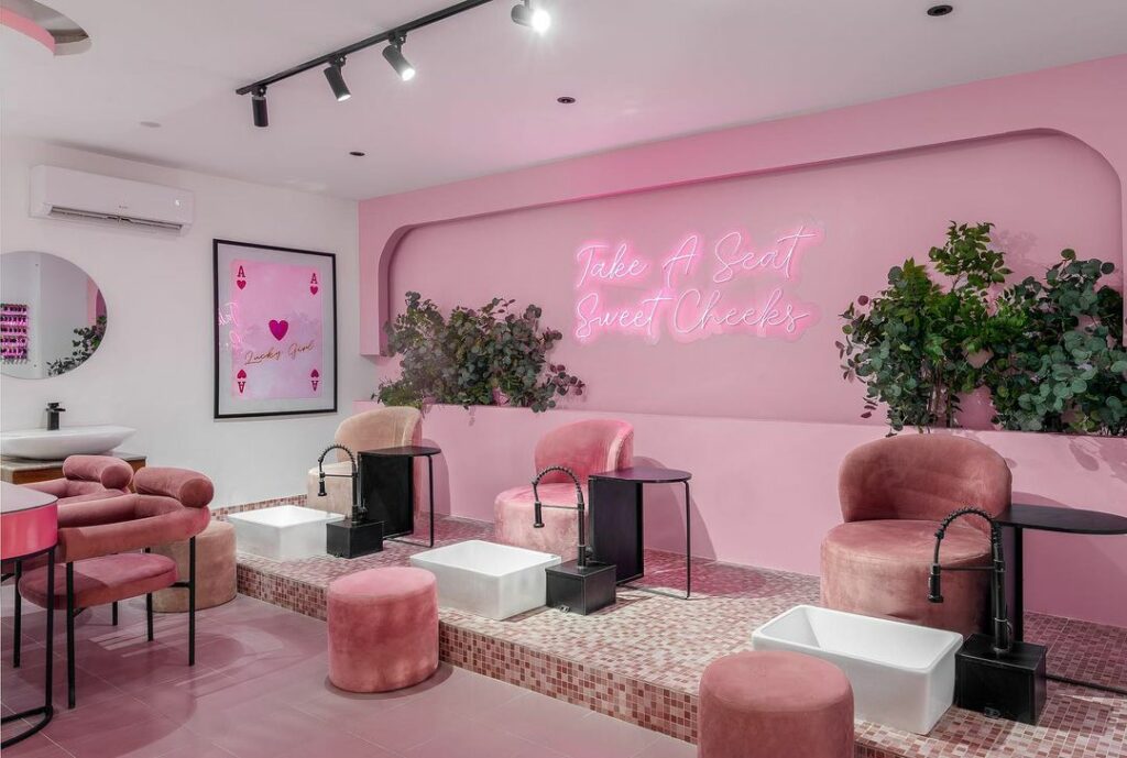 A pink salon interior with a neon sign that reads "Take a Seat Sweet Cheeks". 