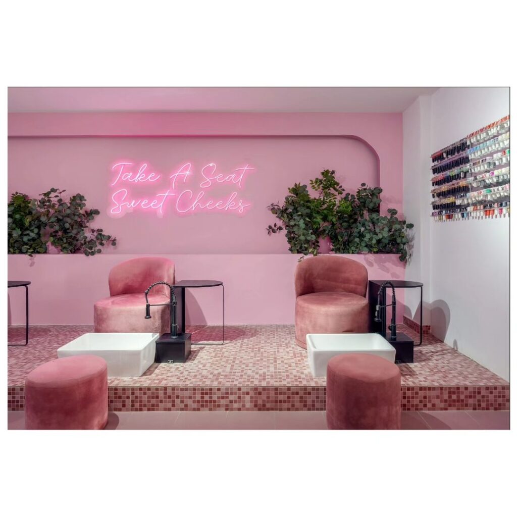 A pink salon with pedicure stations with a neon sign and planting behind.
