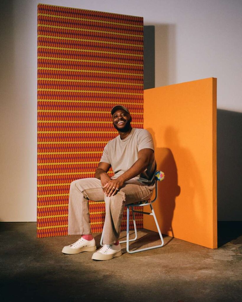 Yinka Ilori sitting on a chair in front of a wall smiling.
