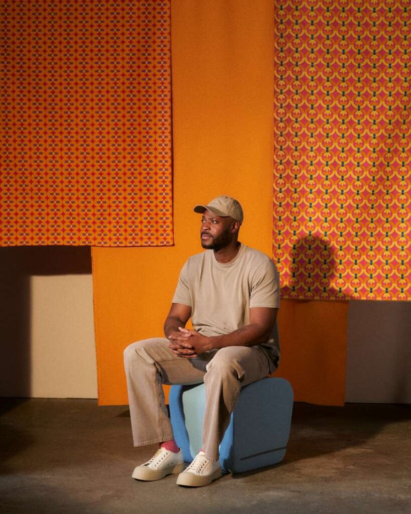 A man sitting on a blue chair in front of a wall covered in vibrant fabric patterns.