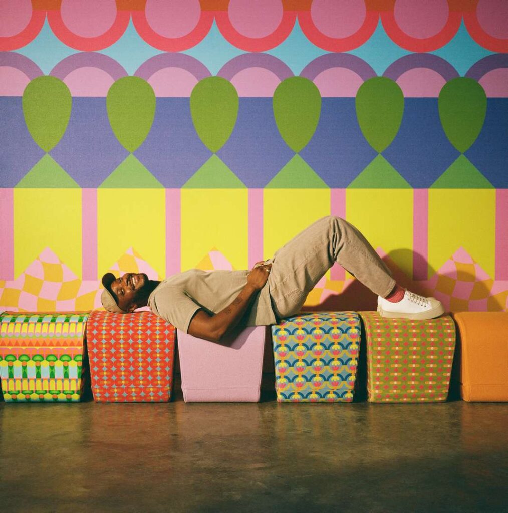 A man lying on colorful seats in front of a vibrant wall, enjoying a sunny day in the park.