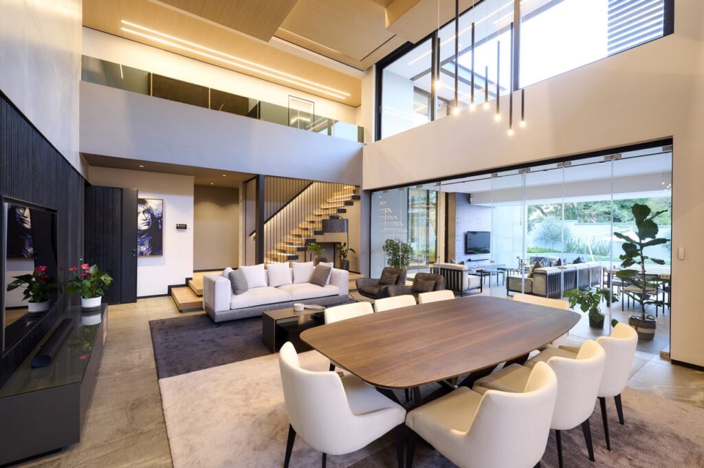 Modern living room with large windows, showcasing a sleek 8 seater dining table and contemporary decor. Also seen are the recessed ceiling linear lights, as well as a chandelier of pendant ligts.