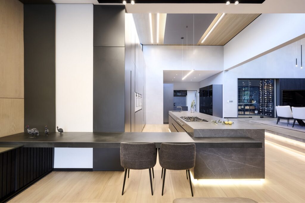 A contemporary kitchen with sleek dark grey countertops, linear pendant lights and warm wooden floors.