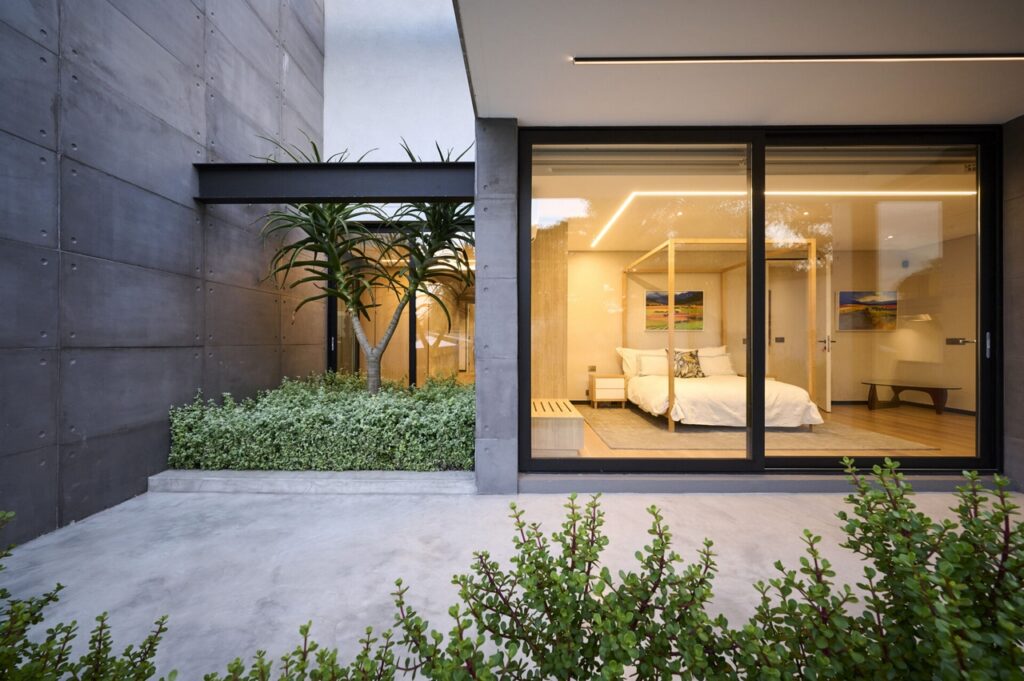 A modern house with concrete exterior finish and a view of the bedroom with sliding glass doors. Outdoor linear light can be seen as well.